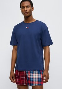 Μπλε ναυτικό βαμβακερό μπλουζάκι με κοντά μανίκια και μικρό λογότυπο στο στήθος, συνδυασμένο με καρό σορτς σε αποχρώσεις κόκκινου, μπλε και λευκού.