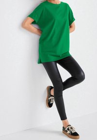 T-shirt verde in cotone con maniche arrotolate, abbinato a leggings neri in finta pelle e sneakers con motivo bianco e nero su uno sfondo chiaro.