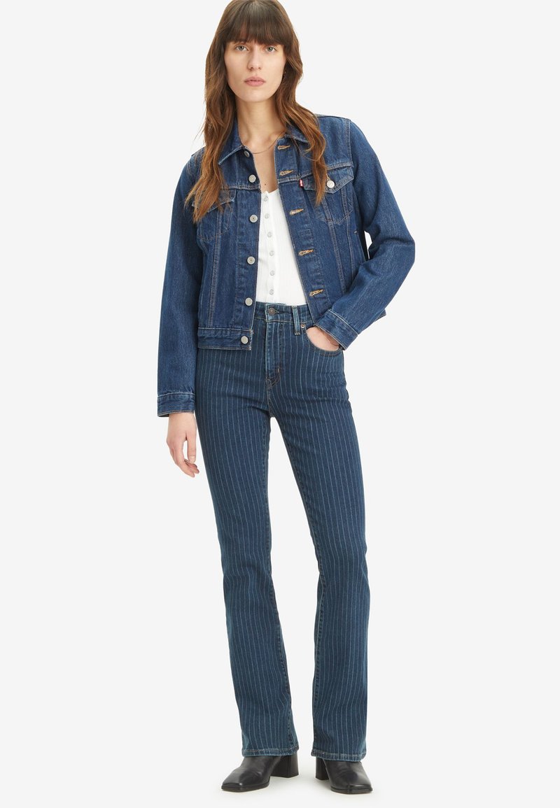 Veste en denim bleu foncé avec un devant à boutons et deux poches poitrine, associée à un jean évasé bleu clair et blanc à rayures et des bottes noires.