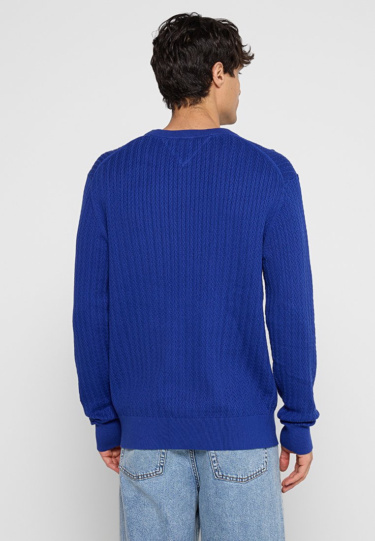 Man in een blauwe kabelgebreide trui en lichtblauwe jeans, staande met zijn rug naar de camera tegen een neutrale achtergrond.