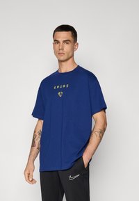 Blå bomulds T-shirt med rund hals, korte ærmer og gul "SPURS" grafik på forsiden. Sammen med sorte bukser med Nike-logo.