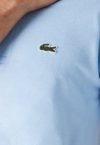 Polo shirt bleu clair en tissu texturé, avec un petit logo de crocodile vert et des accents rouges sur la poitrine gauche.