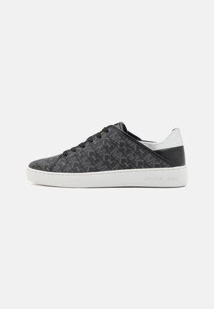 Niedriger schwarzer Sneaker mit subtil gemusterter Textur, weißer Sohle und silbernen sowie schwarzen Details an der Ferse auf einfarbigem Hintergrund.