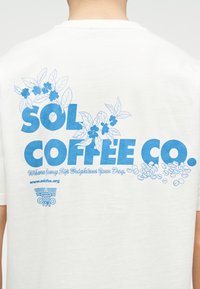T-shirt blanc avec texte bleu "SOL COFFEE CO.", illustrations de plants de café, grains de café, et slogan "Où Chaque Gorgée Éclaire Votre Journée."
