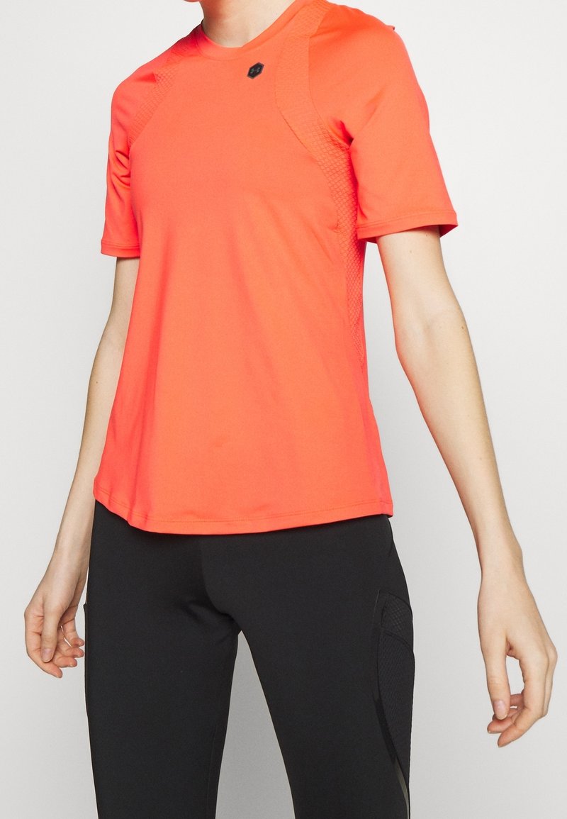 Henkilö, jolla on päällään kirkkaan oranssi lyhytnenäinen urheilupaita ja mustat legginsit, joissa on verkkotasku, seisoo yksinkertaista vaaleaa taustaa vasten.