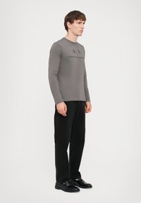 Langermet grå t-skjorte med "Armani Exchange"-logo i svart, sammen med svarte bukser og svarte sko, vist på en mannlig modell.