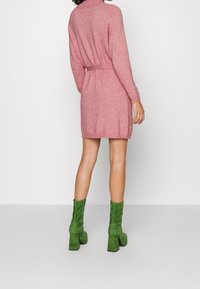 Abito maglione rosa lavorato a maglia con collo alto, vita sottolineata da una cintura e maniche lunghe, abbinato a stivali alla caviglia in suede verde con tacco spesso.