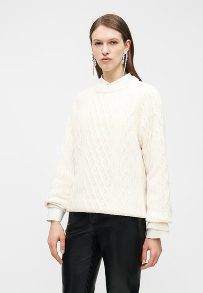 Γυναίκα που φοράει λευκό πλεκτό πουλόβερ με σχέδιο κοτσίδα, μαύρο δερμάτινο παντελόνι και μακριά σκουλαρίκια με κρύσταλλα, στέκεται μπροστά σε έναν απλό φωτεινό φόντο.