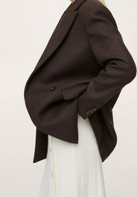 Brown tailored blazer with a structured design, single button closure, side pockets, and textured fabric worn over a white dress.