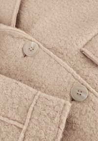 Beige textiel met een zachte, harige uitstraling. Bevat twee ronde, lichtgekleurde knopen en gestikte details aan de randen.