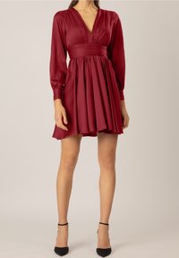 Burgundy klänning med långa ärmar, v-ringning, figurnära överdel och utsvängd kjol, tillverkad av slät tyg. Inkluderar veck och knappdekorationer.