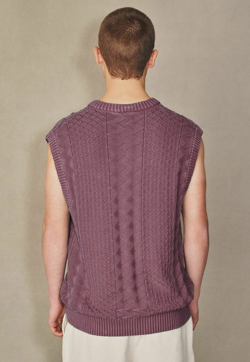 Homme aux cheveux courts portant un gilet en maille violet sans manches et un pantalon clair, vu de dos sur un fond uni.
