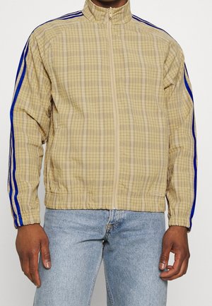 Mann trägt beige karierte Reißverschlussjacke mit blauen Streifen an den Ärmeln und hellblaue Jeans vor weißem Hintergrund.