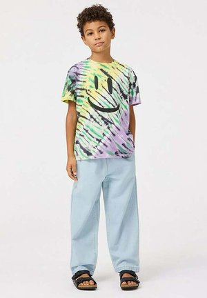 T-shirt w stylu tie-dye w kolorach żółtym, zielonym, fioletowym i czarnym z dużą czarną buźką, w zestawie z jasnoniebieskimi luźnymi spodniami i czarnymi sandałami.
