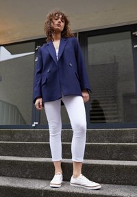 Námořnický dvouřadový blazer s kapsami, nošený přes bílou košili a bílé skinny džíny, doplněný o bílé tenisky na šedých schodech.