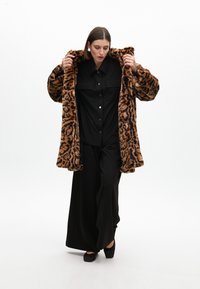 Manteau od umjetnog krzna s uzorkom leoparda, ležernog kroja, uparen s crnom košuljom s dugmadima i crnim hlačama širokih nogavica. Uočljivi detalji teksturirane tkanine.