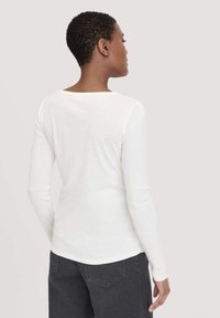 Witte langesleeve top met een bootnek, gladde textuur en een gebogen zoom, gecombineerd met donkergrijze high-waisted jeans.