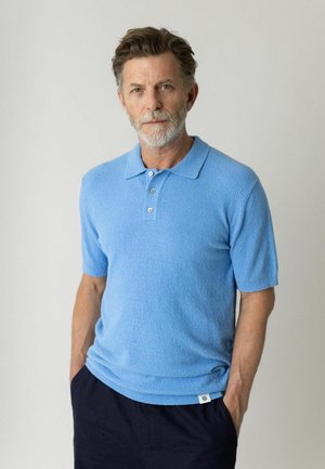 Keskeas vana hallide juuste ja habemega mees, kes kannab helehalli lühikeste varrukatega tekstuuriga polo särki, seisab tumedates pükstes käed taskutes.