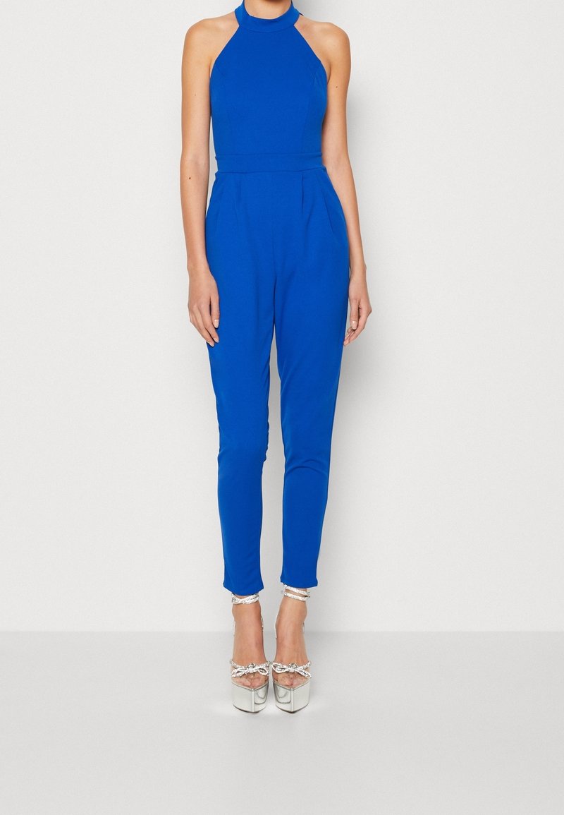 Combinaison sans manches bleu avec un col halter, des jambes fuselées et des plis subtils. Associée à des chaussures à talons hauts métalliques avec des accents en nœud.