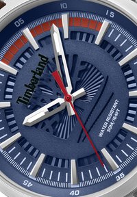 Timberland pulksteņi ar tumši zilu ciparnīcu, sudraba stundu indeksiem, sarkanu sekundes rādītāju un 50m ūdensizturību. Iespējas ietver izteiktu logotipu un rakstainu tekstūru.