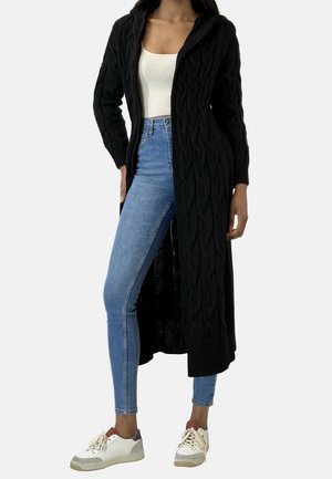 Femme portant un long cardigan noir en maille avec une capuche, un top court blanc, un jean skinny bleu et des baskets blanches sur un fond uni.