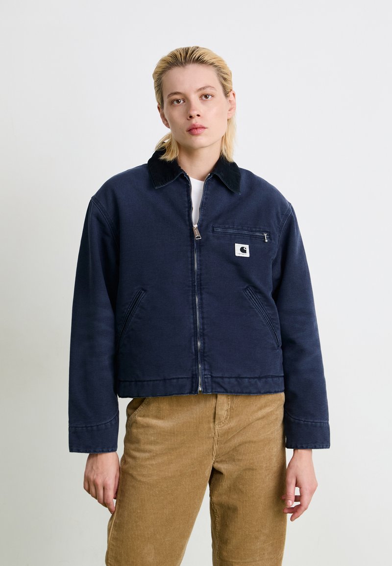 Young person with short blonde hair wearing a navy zip-up jacket with a chest pocket and tan corduroy pants, standing against a plain background.