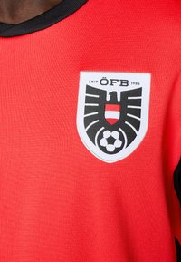 Piros sportmez texturált kivitelben, amelyen egy hangsúlyos fekete-fehér pajzs logó látható focilabda motívummal. Fekete díszítés a galléron.