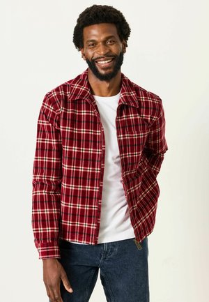 Mann lächelt, trägt eine rote Kariertjacke über einem weißen Hemd und dunkle Jeans vor einem schlichten hellen Hintergrund.