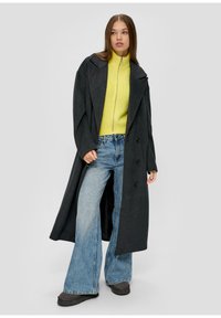Langer grauer Wollmantel mit großen Revers, doppelten Knöpfen; kombiniert mit einem gelben gerippten Pullover und blauen Schlagjeans.