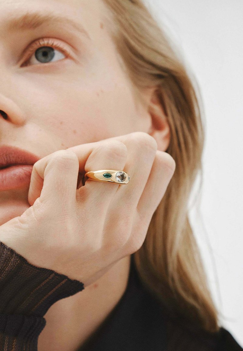 Anillo de oro con una piedra preciosa transparente y un acento verde, colocado en un dedo. Primer plano de una mano situada cerca del rostro contra un fondo neutro.