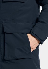 Marinblå vattentät jacka med texturerat tyg, utrustad med en dragkedjeficka, knappad fickklaff och märkesdetalj på bröstet.