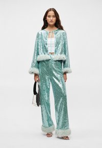 Ένα σετ σε χρώμα τυρκουάζ με παγιέτες που περιλαμβάνει ένα κοντό μπουφάν με ψεύτικη γούνα και ταιριαστά παντελόνια, σε συνδυασμό με ένα λευκό κροπ τοπ και ασημένια τσάντα.