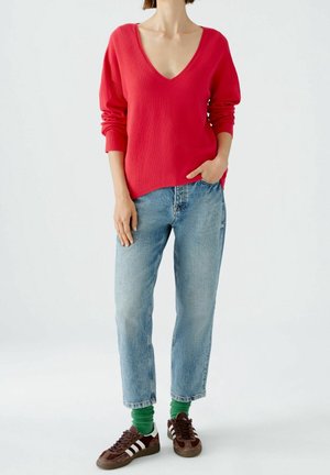 Persoon draagt een felrode V-hals trui, lichtblauwe spijkerbroek, groene sokken en bruine sneakers, staat met één hand in de zak.