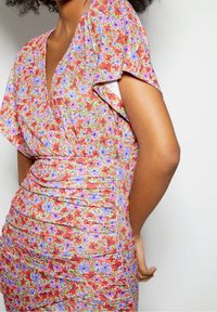 Vestido floral en tonos rojos y morados, con escote en V, mangas acampanadas y textura fruncida a lo largo de la cintura para una silueta ajustada.