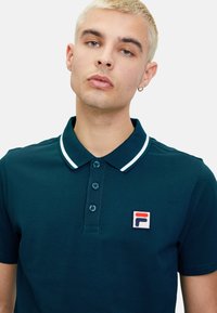 Polo shirt di un verde scuro con colletto, tre bottoni e dettagli bianchi. Presenta una patch quadrata con il logo sul petto sinistro.