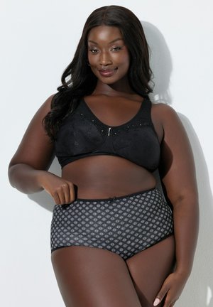 Donna taglia forte con lunghi capelli neri, indossa un reggiseno nero in pizzo e mutandine a vita alta con fantasia, sorridente su uno sfondo bianco.