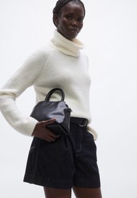 Črna usnjena torbica, ki jo drži oseba, oblečena v kremasti pulover z visokim ovratnikom in temne denim kratke hlače z vidnimi šivi.