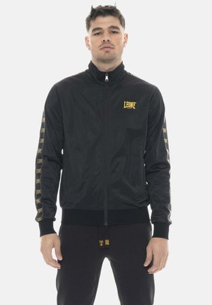 Jonge man met een zwart jack met rits en gele "LEONE"-logo's op de borst en mouwen, staande tegen een witte achtergrond.