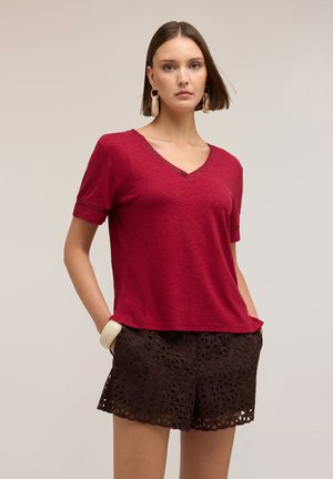 Frau mit schulterlangem Haar, die große goldene Ohrringe, ein rotes V-Ausschnitt-Kurzarmshirt und schwarze Shorts mit Spitzenmuster trägt.