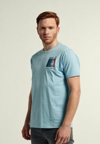 Camiseta de algodón azul claro con mangas cortas, cuello redondo, y gráfico impreso que presenta "POLO" y un diseño en el pecho izquierdo.
