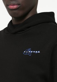 Svart hettegenser med en myk tekstur, med en brodert blå logo på brystet. Designet inkluderer en avslappet halslinje og enkel form.