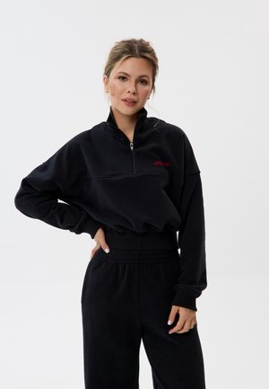 Frau steht mit Hand in der Hüfte, trägt schwarzen Halb-Reißverschluss-Sweatshirt und passende Hose vor weißem Hintergrund.