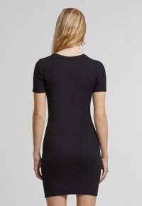 Femme aux cheveux longs et blonds portant une robe noire moulante à manches courtes, tournée de dos sur un fond gris clair uni.