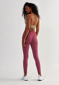 Soutien-gorge de sport beige avec des bretelles croisées, associé à des leggings moulants bordeaux. Tissu texturé avec un logo subtil sur la ceinture.