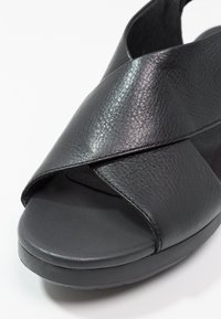 Sandale en cuir noir avec larges lanières croisées et design ouvert à bout rembourré sur une semelle plate.