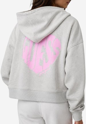 Personne portant un sweat à capuche gris clair et un pantalon, avec un grand logo rose en forme de cœur "GUESS" dans le dos du sweat à capuche.
