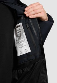 Notranjost črne jakne z mrežasto žepom, tekstilno etiketo in žepom na zadrgo. Vsebuje gladek material in eleganten dizajn.