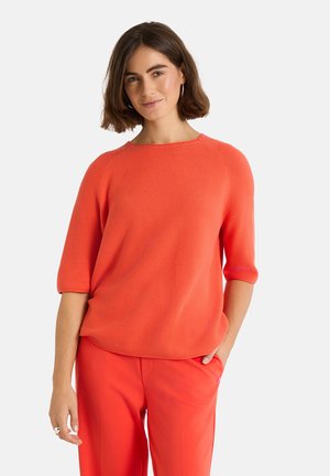 Junge Frau mit kurzen braunen Haaren, die Creolen trägt, einen orangen Strickpullover mit dreiviertel Ärmeln und passende orange Hosen.