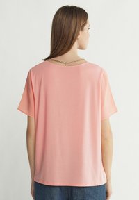 Lösa, kortärmade skjorta i mjuk korallfärgad tyg. Har en rund halsringning med en guldkedjeaccent och en slät textur.