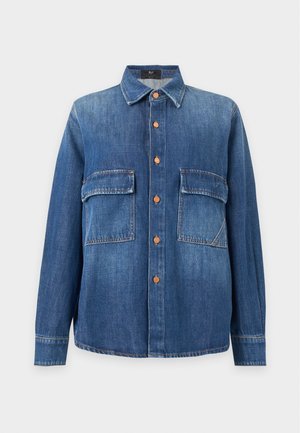 Jeune femme portant un jean en denim bleu foncé ample, un débardeur côtelé beige et une veste en denim bleue déboutonnée, mains dans les poches.